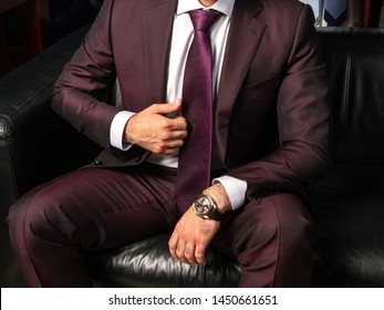 A Man In A Classic Suit Is Sitting On A Black Leather Sofa, Close-up. Men's Suit Italian Designers. Clothing Business Person. Wristwatches Brown. Men's Tie. Spread His Legs