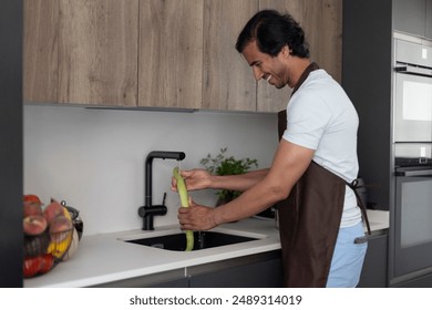 Man cleaning a green vegetable at the kitchen sink, wearing a brown apron, preparing food - Powered by Shutterstock