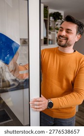 A man is cleaning a kitchen window using a blue cloth. He is smiling while working in a bright kitchen with plants.