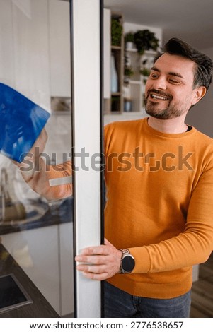 A man is cleaning a kitchen window using a blue cloth. He is smiling while working in a bright kitchen with plants.