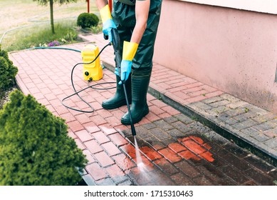 Man cleaning red concrete pavement blocks using high pressure water cleaner. Paving cleaning concept. Man wearing waders, protective waterproof trousers, doing spring jobs in garden.