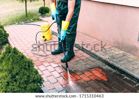 Man cleaning red concrete pavement blocks using high pressure water cleaner. Paving cleaning concept. Man wearing waders, protective waterproof trousers, doing spring jobs in garden.