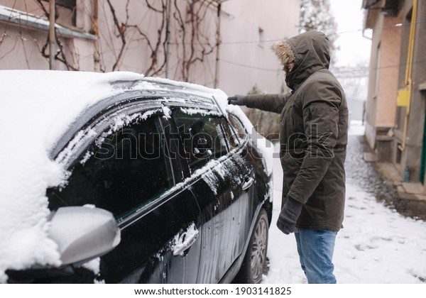 A\
man is cleaning snowy window on a car with snow scraper. Focus on\
the scraper. Cold snowy and frosty morning. Black\
car