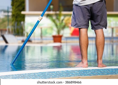 Man cleaning a swimming pool in summer with a brush or net on a blue pole standing barefoot on the tiles at the edge, low angle view of his lower body. - Shutterstock ID 386898466