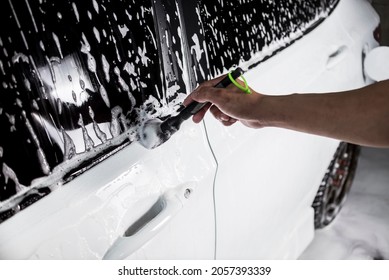 A Man Cleans The Outer Window Seal Of A White Sedan Covered In Car Shampoo With A Fine Brush. A Vehicle Being Serviced At A Carwash Or Auto Detailing Shop.