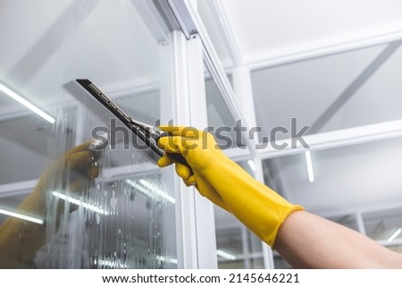 A man cleans the surface of an interior office window with a glass wiper or squeegee. The surface is sprayed with soap.
