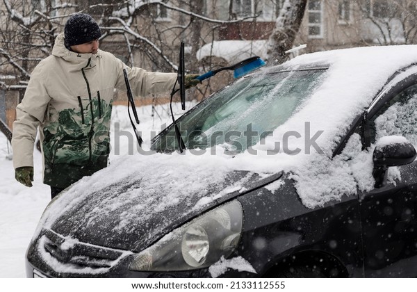 A man\
cleans the windshield of a car from snow and ice with a brush and\
scraper. Cleaning cars from snow in\
winter.