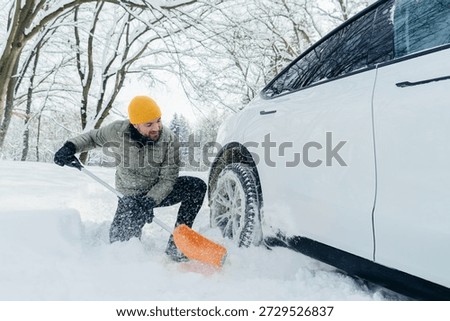Man clearing deep snow blocking a white electric car wheel with an orange shovel during cold winter weather