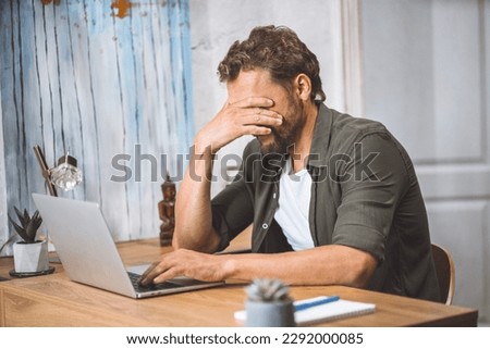 Man clearly overwhelmed and stressed out at work. His head in his hands, covering his face with palm, appearing to be in state of deep depression. Computer in front of him serves as a reminder of the