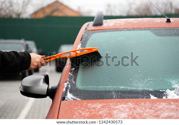 Man clears snow from icy\
windows of car. Brush in mans hand. Windshield of orange auto,\
horizontal view.