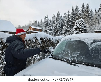 Man clears windshield after snowfall in snowy forest. Snow covers car and nearby hedge. Snowy trees stand behind wooden fence. Overcast sky above rural winter scene