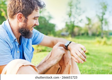 Man clicking on a screen of his smart watch sitting on a grass in a park.  - Powered by Shutterstock