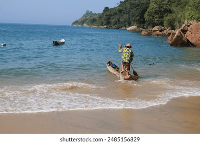 Man climbing into a caiçara log canoe at the edge of the sea with the coast of rocks and vegetation in the background at Praia Grande da Cajaíba, Paraty, Rio de Janeiro, Brazil - Powered by Shutterstock