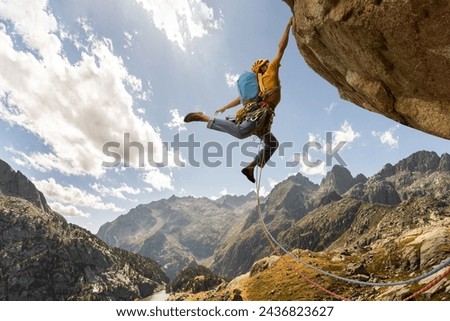 A man is climbing a mountain with a blue backpack. He is wearing a yellow jacket and is jumping off a cliff