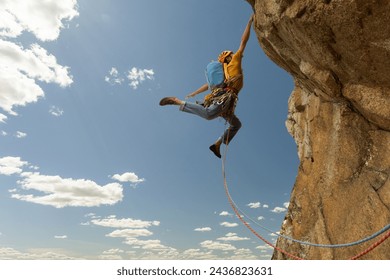 A man is climbing a rock wall with a blue backpack. He is wearing a yellow shirt and is jumping off the wall - Powered by Shutterstock