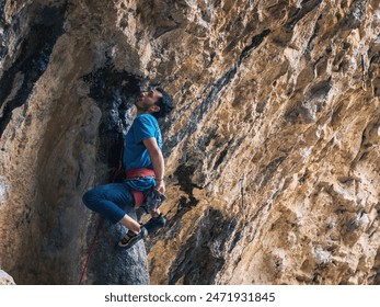 A man is climbing a rock wall. He is wearing a blue shirt and a red climbing harness. He is resting in an acrobatic stance while taking chalk from his magnesium bag. - Powered by Shutterstock