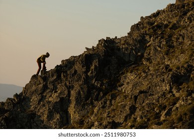 A man is climbing a rocky mountain. The mountain is covered in rocks and the sky is a beautiful shade of blue. The man is wearing a yellow shirt and red pants - Powered by Shutterstock