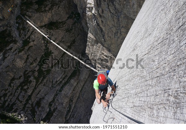 man climbing via ferrata with steps and suspension\
bridge in the background with exposed and challenging passage on\
rock