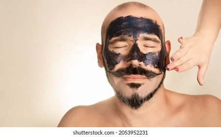 Man With Closed Eyes And Black Face Mask. Woman Puts Exfoliating Scrub On His Face. Bald Adult With Beard And Mustache. Concept Health And Wellness, Skin Care.