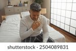 Man clutching chest appears distressed while sitting on bed in bedroom, capturing a moment of potential pain or illness in a home environment.