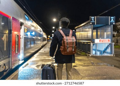 Man in coat and backpack pulls rolling luggage on a dim train platform at night, boarding for a business trip  lone urban traveler heading into the city for work and adventure