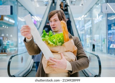 Man in a coat holds a paper check and a bag with a baguette and lettuce in a shopping mall and is shocked by the high prices of groceries.