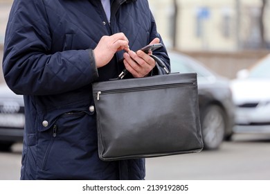 Man In Coat Standing With A Leather Briefcase With Smartphone In Hand On A City Street. Concept Of Businessmen, Official, Government Employee