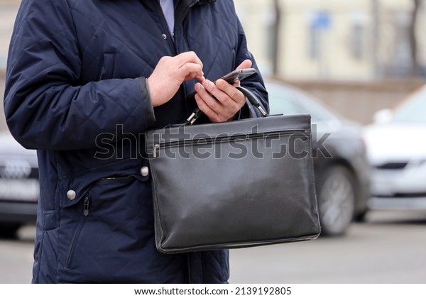 Man in coat standing with a leather briefcase\
with smartphone in hand on a city street. Concept of businessmen,\
official, government\
employee