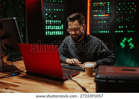 Man coding or hacking on a red laptop in a dark data center, surrounded by busy blinking server rack lights
