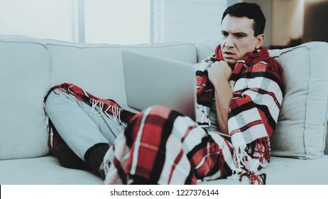 Man With Cold Sitting On Sofa In Checkered Blanket. Man At Home. Sick Young Man. White Sofa In Room. Unhappy Guy. Disease Concept. Healthcare And Healthy Lifestyle Concept. Using Laptop.