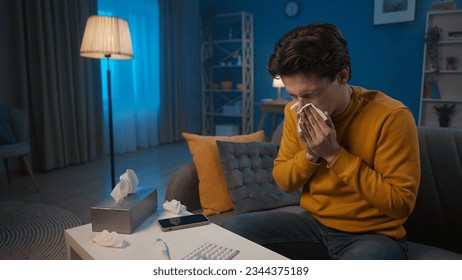A man with cold symptoms is sitting on a sofa in the living room. A man blows his nose into a paper handkerchief. Colds, nasal congestion, allergies. Home medicine concept.