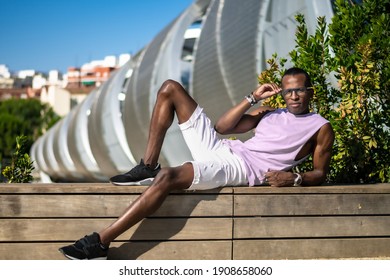 Man of color lies down while holding his glasses, on a wooden bench next to a bridge