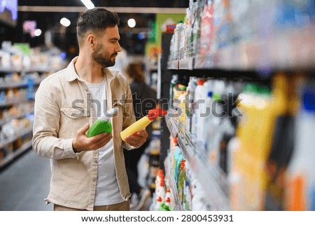 Man comparing two spray bottles of cleaning products in a bright supermarket aisle, making a selection for household needs