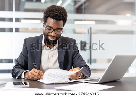 Man completing important documents at his desk in a modern office, concentrating on crucial details and finalising agreement with a pen while laptop and smartphone are nearby