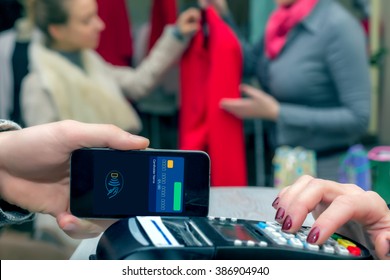 Man Completing Mobile Payment Using Modern Smart Phone Technology At Store Cashiers Desk With Terminal Salesperson And Female Customer Interacting On Background