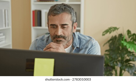 Man at computer with beard looking up, hands clasped under chin, wearing denim jacket, monitor with yellow sticky note, bookshelf and plant in studio; thoughtful focus concentration.