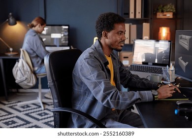 Man concentrating on computer with tablet and coffee while woman in background working on desktop. Office filled with modern technology and organized workspace - Powered by Shutterstock