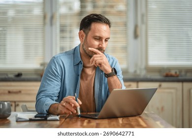 Man is concentrating while working remotely from a domestic setting, pondering a solution and carefully reviewing information on his laptop screen with a pen in hand