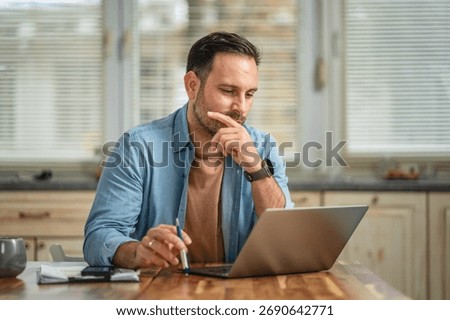 Man is concentrating while working remotely from a domestic setting, pondering a solution and carefully reviewing information on his laptop screen with a pen in hand