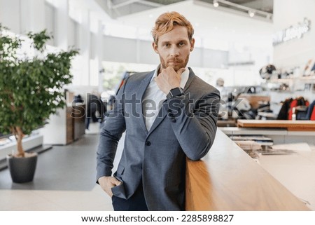 Man confident serious successful businessman caucasian male client 20s in classic grey suit prop up chin looking camera standing at office table in dealership store indoors Business lifestyle concept.