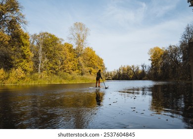 A man confidently and steadily balances atop a paddle board located in the tranquil center of a serene lake, which is magnificently embraced by natures stunning and picturesque landscape around it - Powered by Shutterstock