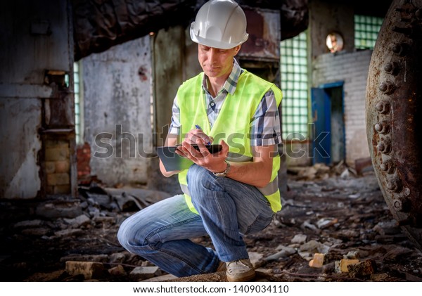 A man in a construction\
helmet and with a black folder in his hands at a construction site.\
Brigadier in working form, inspection of the structure. A man takes\
notes.