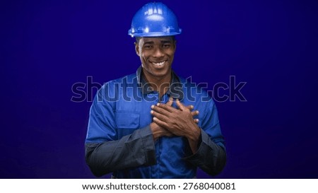 Man construction worker in blue hardhat and coveralls holding hands to chest while smiling in studio; gratitude dedication.