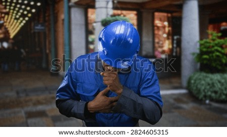 Man construction worker in blue hardhat and blue work uniform coughing into fist and clutching chest on street near shopfronts; medical distress.