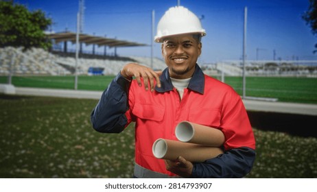Man construction worker in hard hat and red safety jacket holding rolled blueprints and making ok sign while standing by stadium field in building; confidence planning safety.