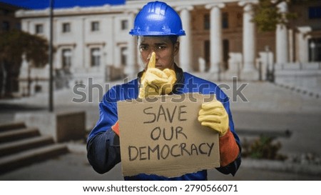 Man construction worker in hardhat holds cardboard sign save our democracy and places finger to lips for silence in front of government building; urgent plea.