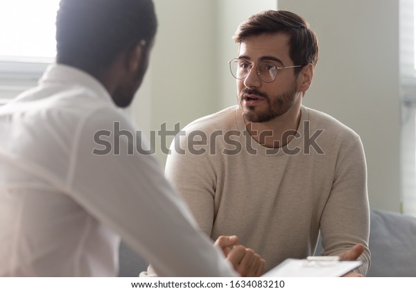 Man consulting with practitioner, African\
psychologist holding clipboard with card sitting in front of\
patient listens his mental health complaints. Job interview process\
applicant and HR manager