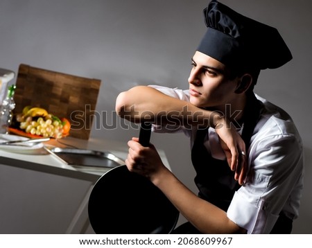 Man cook is thinking about something. He is holding a frying pan in his hands. Portrait of a cook in kitchen. Young chef is preoccupied with something. Chef of restaurant is perplexed.