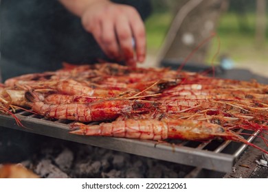 A Man Cooks Large Shrimp Langoustines On An Open Fire On The Grill With Smoke. BBQ Outdoor. Grilled Sea Food Outdoor Party Picnic In The Park. Close Up.