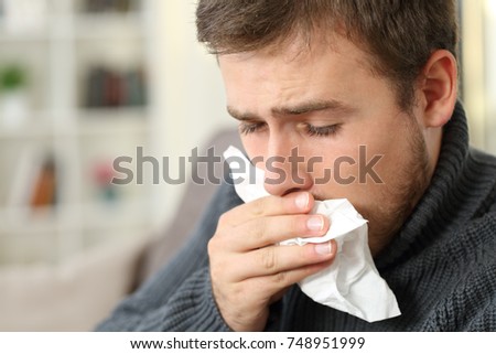 Man coughing covering mouth with a tissue sitting on a couch in a house interior